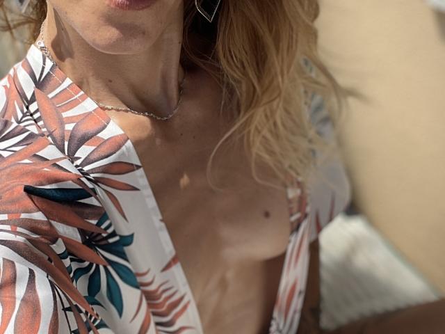 Het topje van DaneeSoHot is open, waardoor een bruine streep en kleine borsten zichtbaar zijn. Ze draagt een bloemenprintshirt, heeft golvend blond haar en een zilveren ketting, op een licht onscherpe foto..