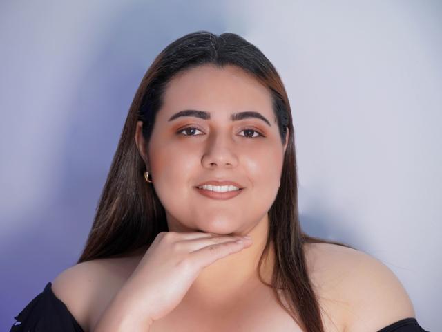 VeloraBliss, con capelli castani, pelle abbronzata e seno piccolo, sorridente, con una mano sotto il mento, indossa una maglietta nera con una spalla scoperta, orecchini dorati, luce soffusa, espressione rilassata..