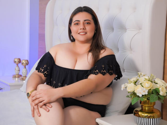 VeloraBliss, een vrouw met een grotere maat en donkerbruin haar, zit op een witte, gestoffeerde stoel, gekleed in een zwart top met een open schouder en zwarte onderbroek, waarbij haar grote borsten zichtbaar zijn..