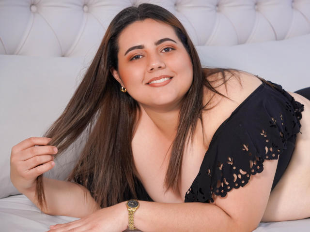 VeloraBliss, una donna formosa con lunghi capelli castani, sorride mentre è sdraiata su un letto, indossa una maglia nera con una spalla scoperta, tiene i capelli, mostra il seno e porta un orologio d'oro..