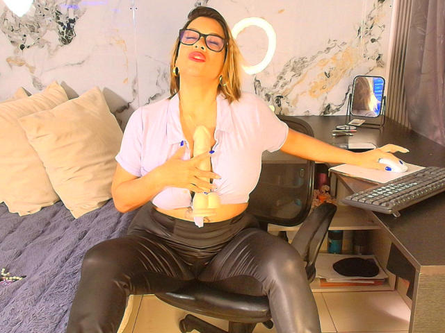 AlixLow with glasses, pink shirt, and black leather pants, using a fleshlight on her large tits while sitting in a chair, in front of a desk with a ring light.