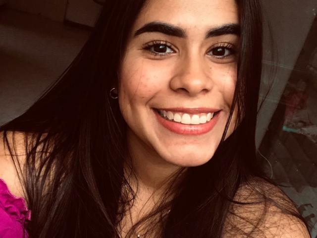 Alicya Torres hat langes, schwarzes Haar, dicke Augenbrauen, braune Augen und ein breites, lächelndes Gesicht mit rosafarbenen Lippen und kleinen, runden, weißen Zähnen. Sie trägt ein rosa Oberteil mit Rüschen..