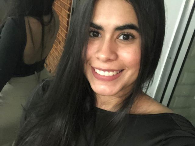 Alicya Torres tiene el pelo largo y negro, ojos marrones y una gran sonrisa que muestra sus dientes blancos. Lleva una camisa negra, y su reflejo es ligeramente visible en un espejo..