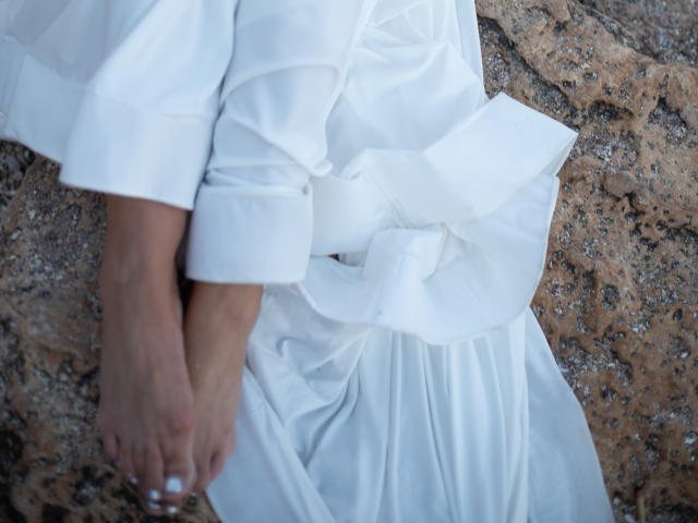 TianaQLove's white dress is bunched at the waist, exposing her bare feet and hands, against a rocky surface. Her arms are folded, showing a relaxed, casual pose.
