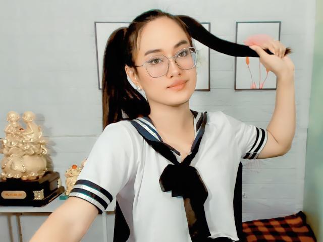 HolyCuteAngel with pigtails, glasses, and a sailor uniform, holding a black hair strand, showing a slight smile, fair skin, and small tits, in a bright room.