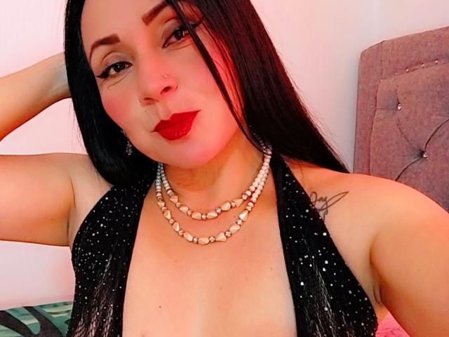 Zaria69 with long black hair, red lipstick, and a nose ring, wearing a black sequined halter top and pearl necklaces, showing her tits, posing with one arm behind her head.