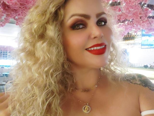 AnnaRichie, con i capelli biondi ricci, il rossetto rosso e una collana, sorride alla fotocamera, mostrando il seno. Sullo sfondo si vedono rami di ciliegio in fiore rosa..