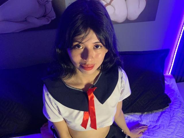 LydiaWels with dark hair, wearing a sailor-style top with a red ribbon, sitting on a bed, purple light in the background, looking up with a slight smile.
