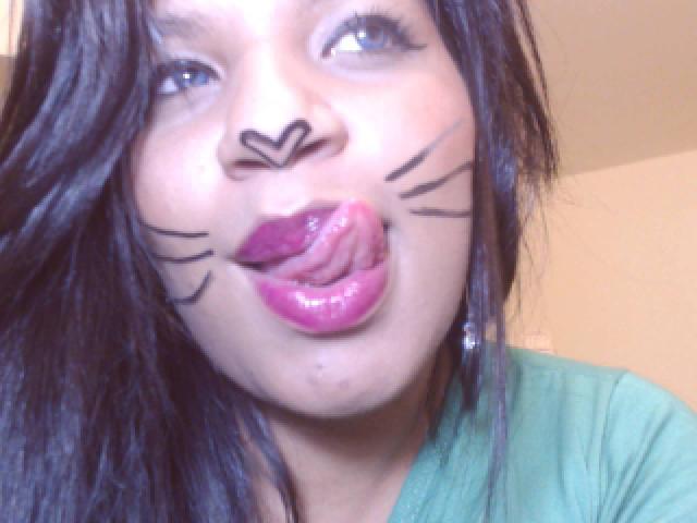 TiaraKitty has black cat makeup on her face, pink glossy lips, and is sticking out her tongue. She has blue eyes, long black hair, and is wearing a green shirt.