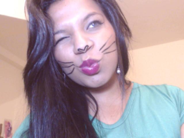TiaraKitty with dark brown hair, winking, black whisker makeup, purple lipstick, green shirt, and pearl earrings, puckering her lips.