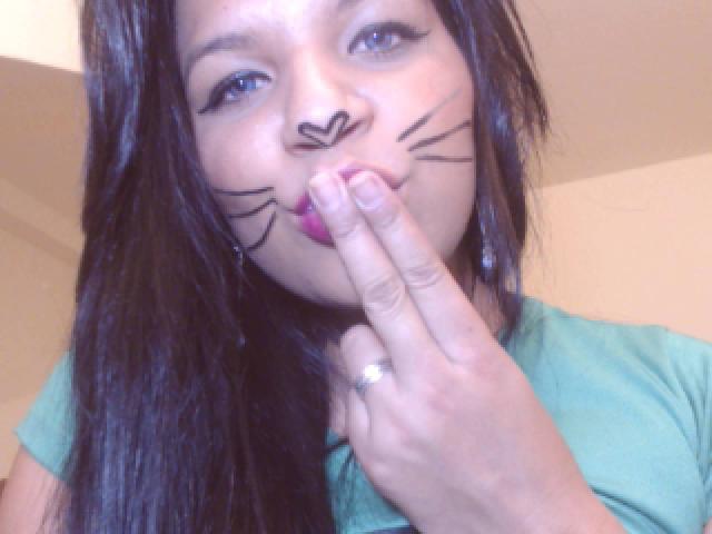 TiaraKitty with dark hair, green shirt, and black face paint resembling whiskers and a heart, holding finger to pink lips, wearing a ring.