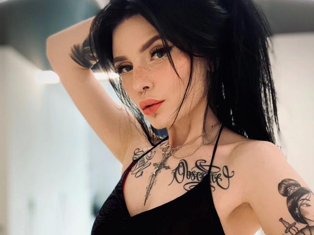 Coco1996-hot with black hair, nose ring, and tattoos, wearing a black halter top, raising arm, showing freckles, red lips, and a necklace, in a bright room.
