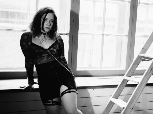 NanaMoonlight with dark, wavy hair, wearing a camo top, black shorts, and a chain necklace, leaning against a window, standing next to a ladder, black and white.