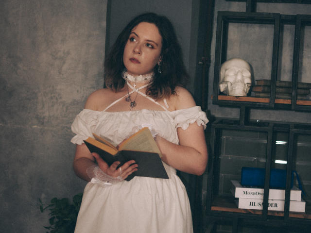 NanaMoonlight with curly dark hair, white off-shoulder dress, holding books, wearing lace choker, skull on shelf, looking to the side, in dimly lit room. Boobs partially visible.