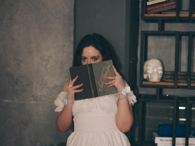 NanaMoonlight with dark hair, wearing a white dress, holding a book, covering her tits with her hands, standing next to a shelf with a white skull.
