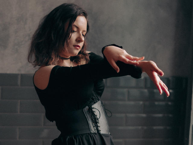 NanaMoonlight with wavy dark hair, wearing a black off-shoulder top and corset, raises her arms gracefully, showing off her tits and delicate fingers in a dark room.