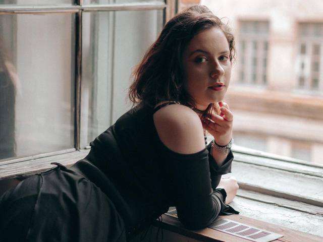 NanaMoonlight with wavy brown hair, black off-shoulder top, and black skirt, leans on a window, looking back with a slight smile, fingers touching her chin.