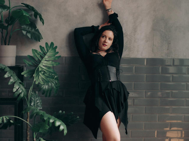 NanaMoonlight with black dress, corset, and thigh-high slit, leaning against wall, arm raised, green plant to left, gray brick wall, slightly seductive, dark hair, subtle smile.