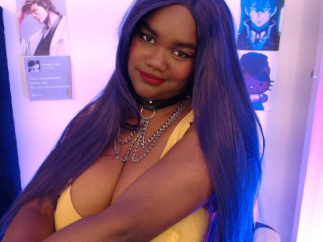 RoseBeckers with purple hair, black choker, and chains, wearing a yellow top showing her big tits, smiling at the camera. She's got dark skin and red lipstick.