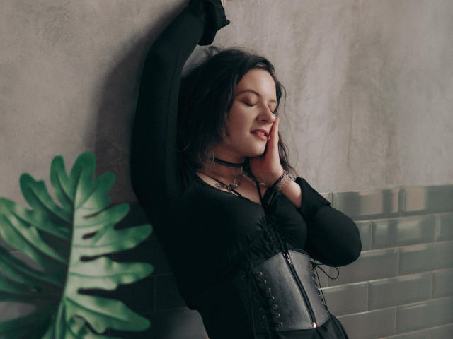 NanaMoonlight with her eyes closed, black dress, and corset, arms raised, hand on cheek, green plant in the foreground, wearing a choker and bracelet.