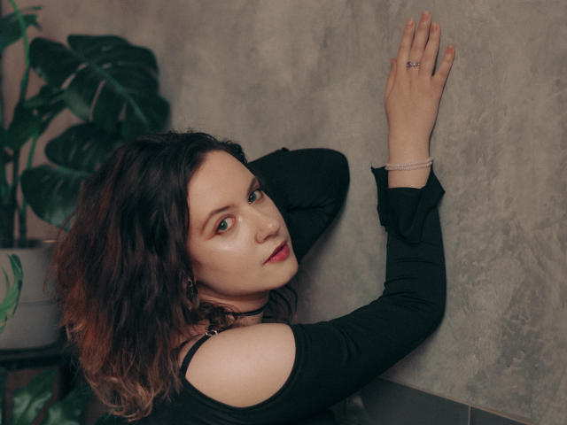 NanaMoonlight with wavy brown hair, green eyes, and red lipstick, wearing a black off-shoulder top, arms against a wall, looking over shoulder, ring on finger.