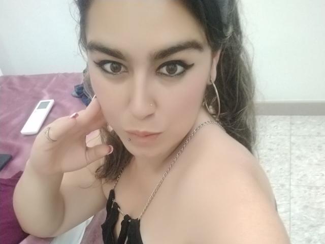 TaylorNova with wet brown hair, pouting lips, wearing a black top that exposes her small, round tits and dark nipples, looking down at the camera.