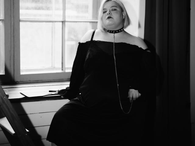 Schwarz-weißes Foto von EllaBigBabe mit heller Haut, kurzen blonden Haaren, die ein schwarzes Kleid mit Schulterauschnitt, eine stachelige Halskette und eine Kette trägt, sitzend am Fenster und nach oben schauend..