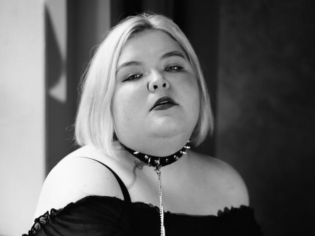 EllaBigBabe, una donna robusta con i capelli biondi corti, è seduta vicino a una finestra, indossa un abito nero con una spalla scoperta e una collana a punte, e tiene una catena contro il suo seno..