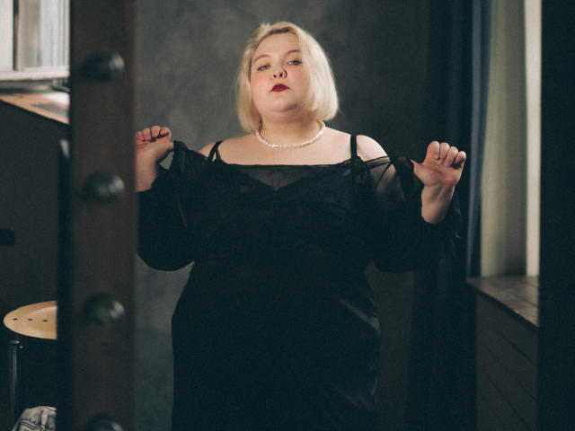 Foto em preto e branco de EllaBigBabe com cabelo loiro curto, vestindo um vestido preto com um ombro descoberto, um colar com spikes e uma corrente, sentada perto de uma janela, olhando para cima com batom escuro..