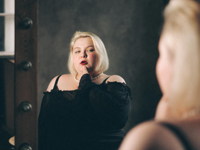 EllaBigBabe stands in a dark room, pulling down black sheer sleeves of a dress, showing her big tits, wearing a pearl necklace, with short blonde hair and red lipstick.