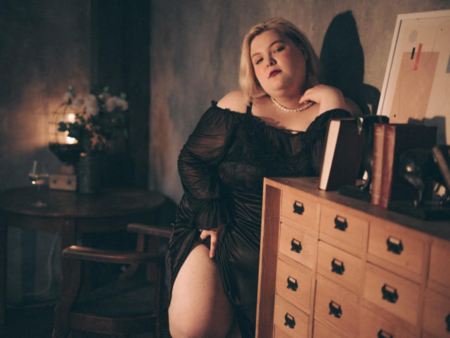 EllaBigBabe with blonde hair, wearing a black, off-shoulder dress, pearl necklace, and red lipstick, leaning on a wooden dresser with books, looking sultry. Her tits are visible.