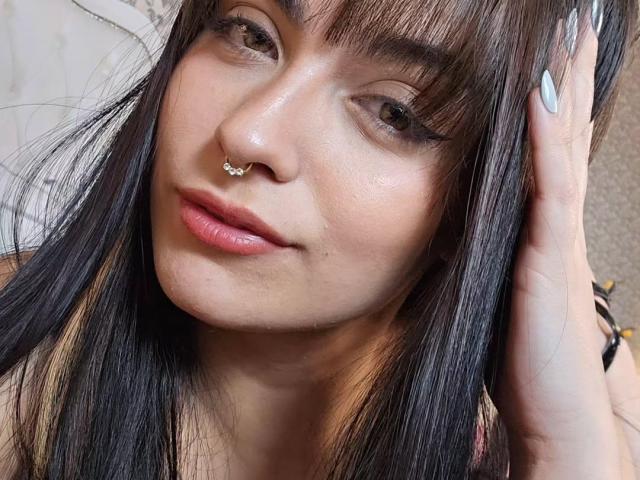 Charlotte Aristizabal mit langen, braunen Haaren, die ein weißes Korsett und blaue Jeans trägt, ihre Brüste hält, Tätowierungen am Arm hat, vor einem Hintergrund mit grünen Ranken steht und mit pinkem Lippenstift in die Kamera schaut..