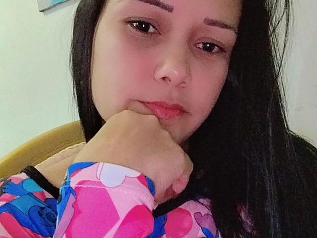 MichellSophia with long black hair, pinkish skin, and brown eyes, resting her chin on her hand, wearing a colorful heart-patterned shirt, looking slightly bored.