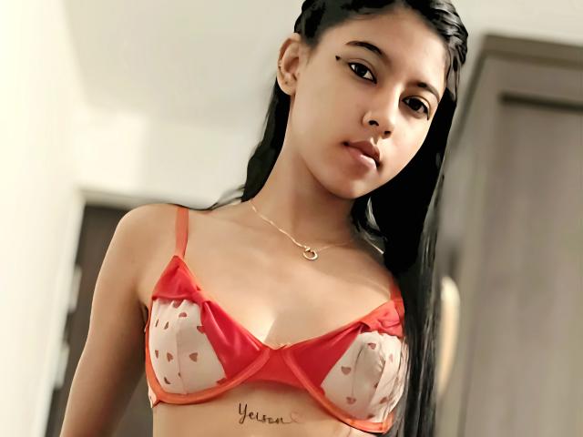 StrawberryAndJennifer with long black hair, wearing a red bra with white heart patterns, showing her small tits, and a tattoo under her tits that reads "Yellow".