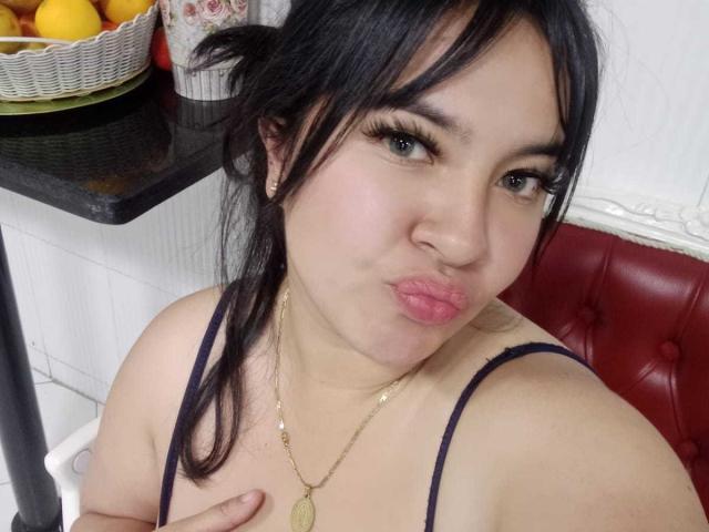 SofiaRutso with pouty lips, black hair, and eyeliner, wearing a black spaghetti strap top, gold necklace, touching her small tits, and giving a cute, sexy look.