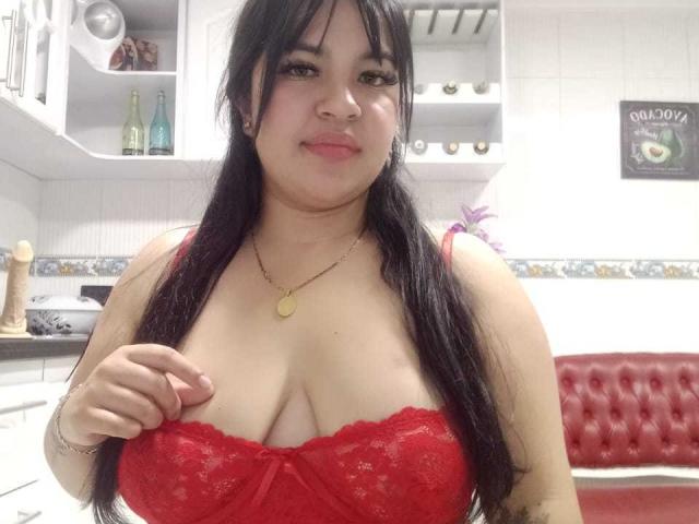 SofiaRutso with long black hair, wearing a red lace bra, pulling down to expose her big tits, smiling, in a kitchen with white cabinets and red tufted bench.