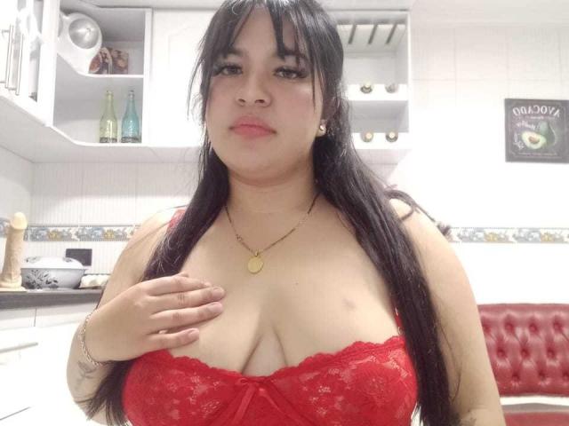 SofiaRutso with long black hair, wearing a red lace bra that shows her big tits, a gold necklace, and a slight smile, standing in a kitchen.