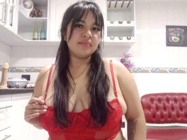 SofiaRutso, chubby Latina with long black hair, red lace bra, showing her tits, smiling, hand on bra, in white kitchen with red tufted chair.