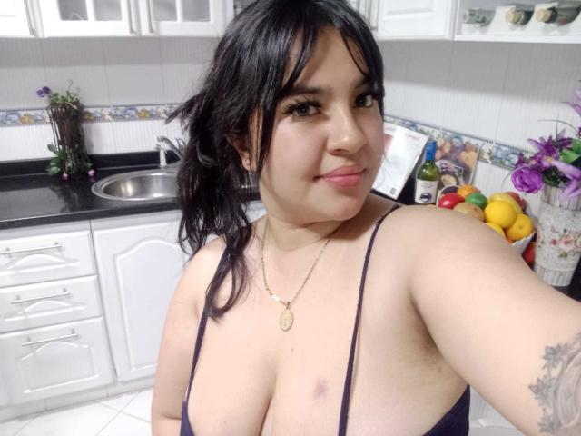 SofiaRutso with dark hair, medium skin, wearing a low-cut purple top, showing large tits, slight bruise on right tit, tattoo on left arm, smiling in a kitchen.