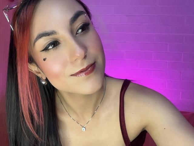 AmyStreetwise69 has pale skin, black and red dyed hair, eyeliner, black heart on cheek, nose ring, glossy pink lips, silver necklace, and is smiling at the camera.