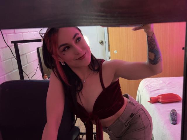 AmyStreetwise69, with red and black hair, medium tits in a red velvet bra, tattooed arm, and gray pants, leans under a table, smiling, in a dimly lit room.