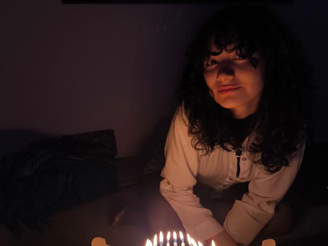 AmeliaTorres with curly dark hair, white shirt, and small tits, kneeling by candlelit table, smiling warmly, hands on candles, dimly lit, shadowed face, cozy nighttime vibe.