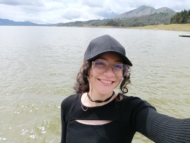 AmeliaTorres with curly brown hair, black cap, round glasses, black choker, and black cut-out top, smiling at the camera, standing in shallow green water, mountain background.