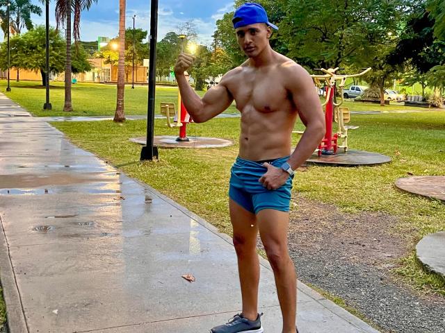 CarlosSmith, shirtless, wearing blue shorts and a blue cap, flexing right arm, standing on wet sidewalk, tree in background, grass, playground equipment, slight tattoo on right thigh.