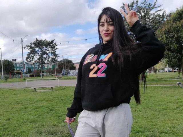 AnalisseJade with long black hair, wearing a black hoodie and gray pants, making a peace sign with her right hand, standing on a grassy area.