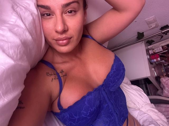 CheyenneSinLimites in blue lace lingerie, showing her tits and a small tattoo on her right shoulder, lying on white bedding with a relaxed, slightly smug expression.