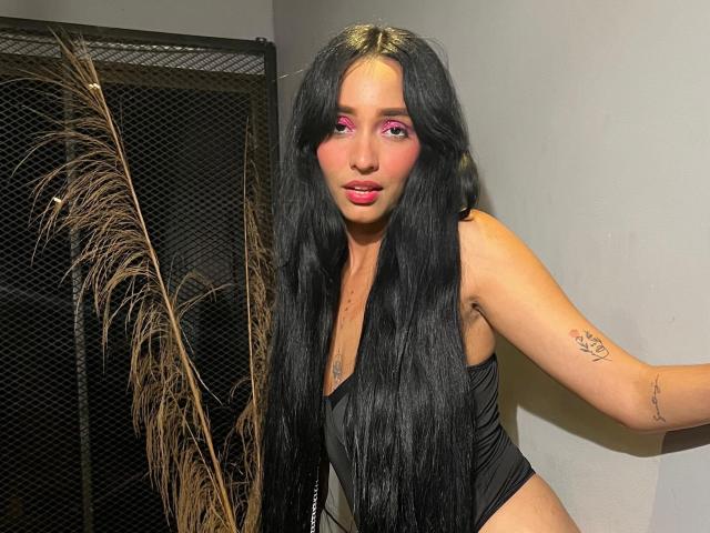 AuraSeductress with long black hair, pink eye makeup, black tank top, visible tattoo on right arm, standing with pouty pink lips, leaning against wall, holding feather.