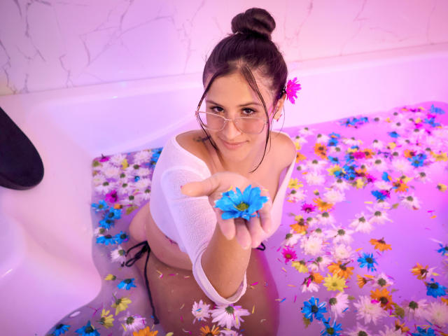 PeytonGrayy dans une baignoire remplie d'eau et de fleurs colorées, tenant une fleur bleue dans sa main droite, portant des lunettes et un haut blanc à une épaule, regardant vers le haut..