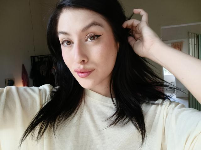 IsabellaPalacio with black hair, light skin, winged eyeliner, pink lipstick, hand in hair, white shirt, looking at camera, slight smile, soft natural light, casual selfie.