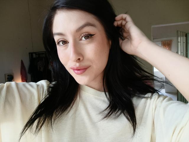 IsabellaPalacio with pale skin, dark straight hair, and pink lips, wearing a cream shirt, hand in hair, subtle eyeliner, slight smile, brown eyes, soft natural light.
